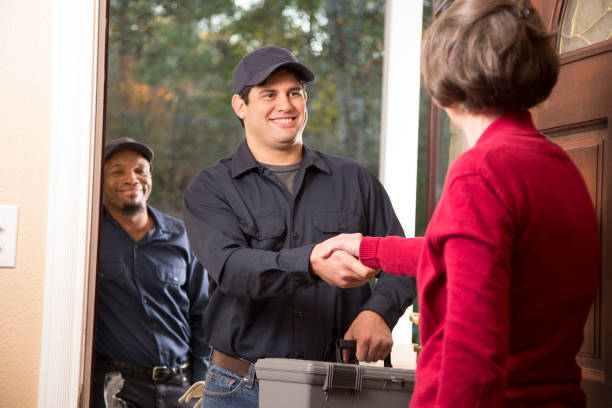 Multi-ethnic team of repairmen arrive at customer's front door to make requested home repairs. Female customer opens door to greet team. Men wear blue uniforms and carry equipment. Focus on man center shaking hands with woman.