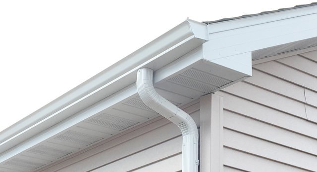 This image shows the exterior corner of a house featuring white vinyl siding, white gutters with a downspout, and white soffit and fascia trim work under the roofline.
